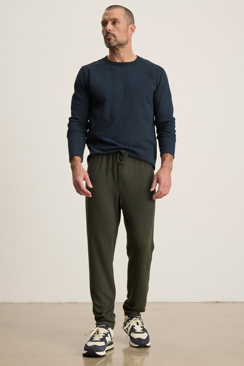 A man stands indoors against a plain background, wearing a navy long-sleeve shirt, black-and-white sneakers, and Velvet by Graham & Spencer’s ZEALAND SWEATPANT in olive green with an elasticized drawstring waist.