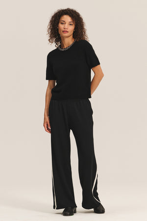 A woman wears the BRIANA SWEATER by Velvet by Graham & Spencer paired with loose black pants with white stripes and black shoes, set against a plain light background—ideal inspiration for your cold-weather wardrobe refresh.