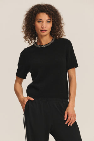 A woman with curly hair wears the Velvet by Graham & Spencer BRIANA SWEATER and black pants, standing against a plain light background.