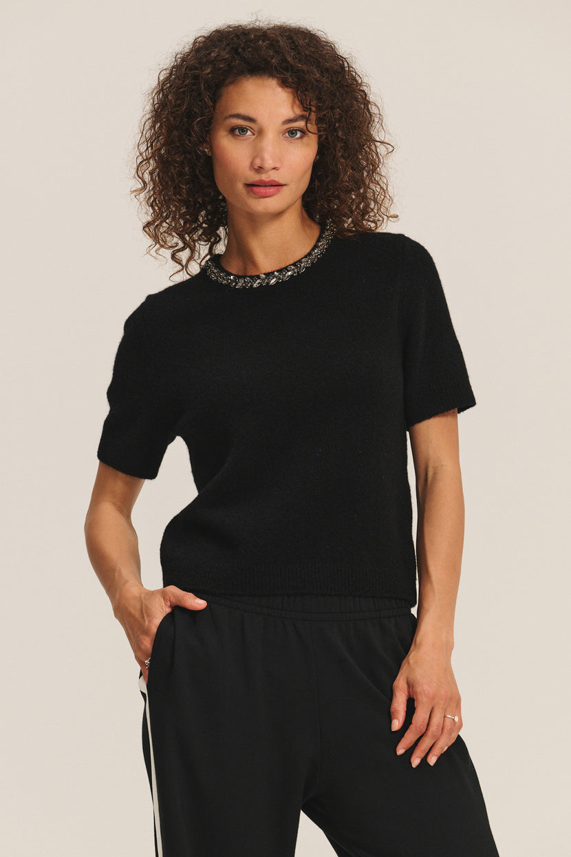 A woman with curly hair wears the Velvet by Graham & Spencer BRIANA SWEATER and black pants, standing against a plain light background.