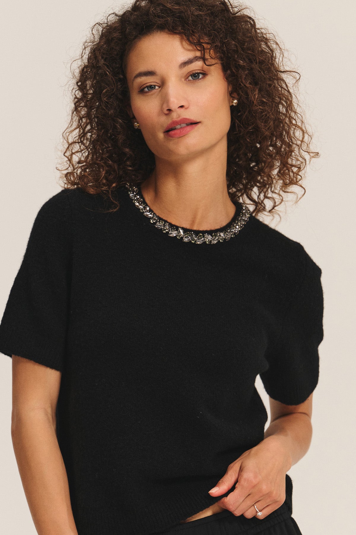 A woman with curly hair models the BRIANA SWEATER by Velvet by Graham & Spencer, featuring short sleeves and a jeweled neckline—a chic cold-weather essential—against a plain light background.-40263647690945