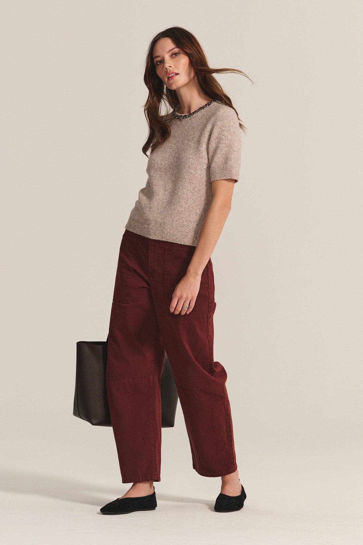 A woman stands against a plain background wearing the BRIANA SWEATER by Velvet by Graham & Spencer—a beige wool blend piece—paired with loose burgundy pants, black flats, and a large black bag for chic cold-weather style.-40263647658177