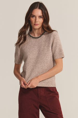 A woman with long brown hair wears the BRIANA SWEATER by Velvet by Graham & Spencer, a short-sleeve wool blend with a beaded neckline, paired with burgundy pants against a plain light background.