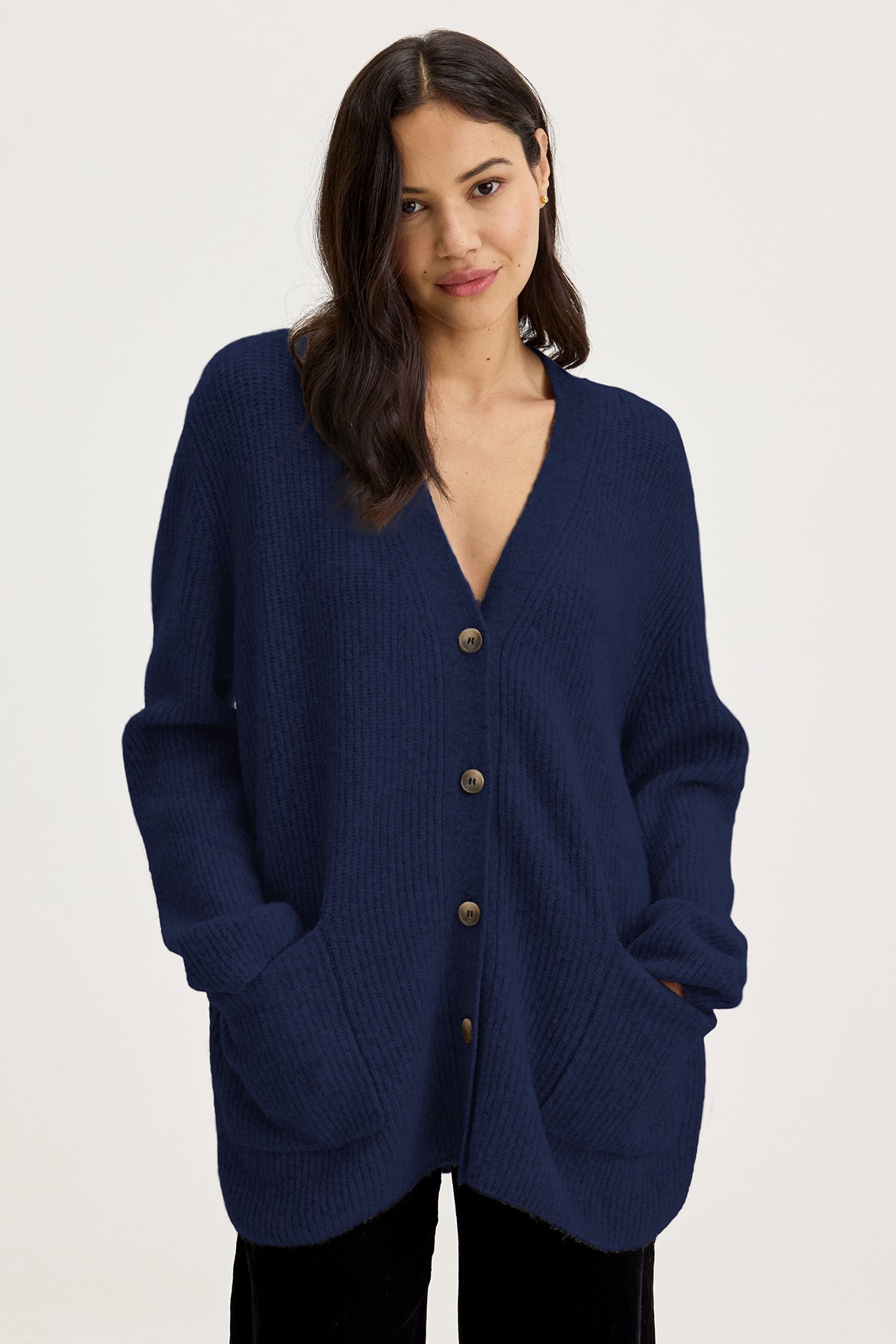   Person standing wearing the Velvet by Graham & Spencer BRITT CARDIGAN, a blue, long-sleeved, wool blend buttoned cardigan with hands in pockets, perfect for layering in cold weather. 