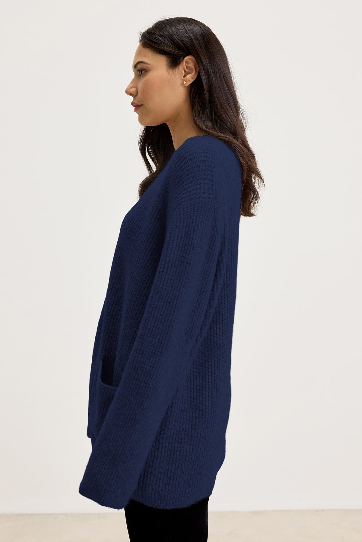   A woman with long dark hair is wearing the BRITT CARDIGAN by Velvet by Graham & Spencer, a dark blue, long-sleeved wool blend sweater, paired with black pants. She is standing in profile against a plain white background, ready for cold weather layering. 