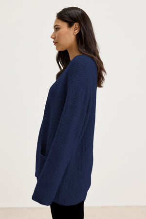 A woman with long dark hair is wearing the BRITT CARDIGAN by Velvet by Graham & Spencer, a dark blue, long-sleeved wool blend sweater, paired with black pants. She is standing in profile against a plain white background, ready for cold weather layering.