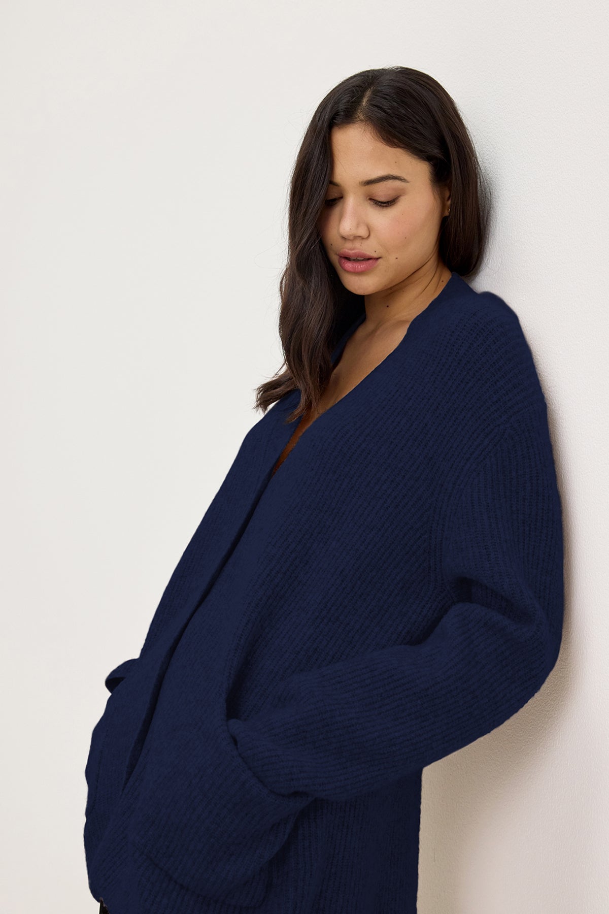   A person with long dark hair is standing against a light wall wearing the Velvet by Graham & Spencer BRITT CARDIGAN in dark blue, with hands in pockets, looking down. 