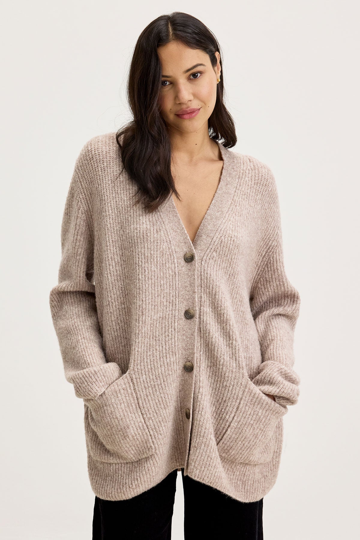 A person with long hair is posing while wearing the BRITT CARDIGAN, a beige wool blend knit cardigan by Velvet by Graham & Spencer, which features front pockets. Paired with black pants and standing against a plain background, this outfit is perfect for layering during cold weather.-37856069157057