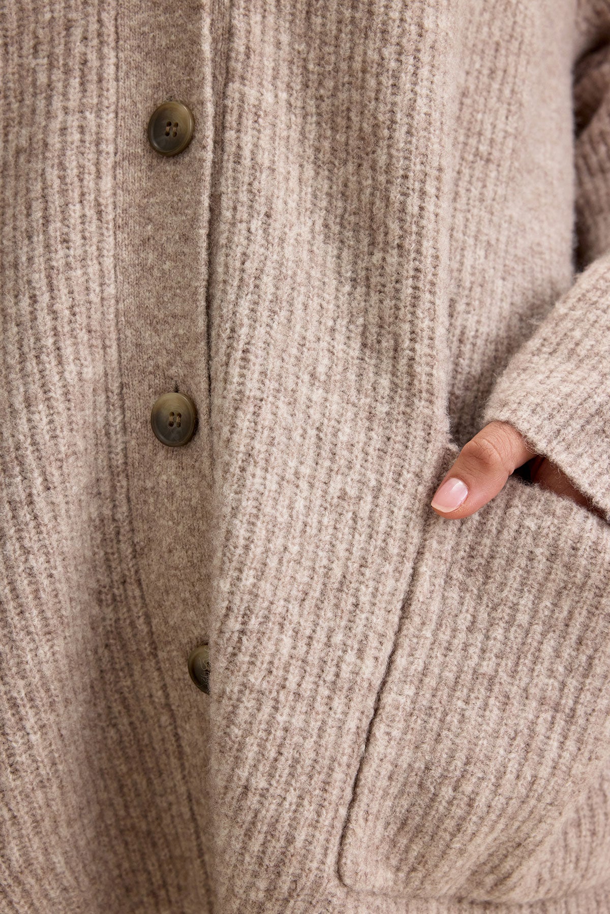   A close-up of a person wearing the BRITT CARDIGAN by Velvet by Graham & Spencer, featuring a beige, rib-knit wool blend with large buttons. Ideal for layering in cold weather, the person is placing one hand into a side pocket. 