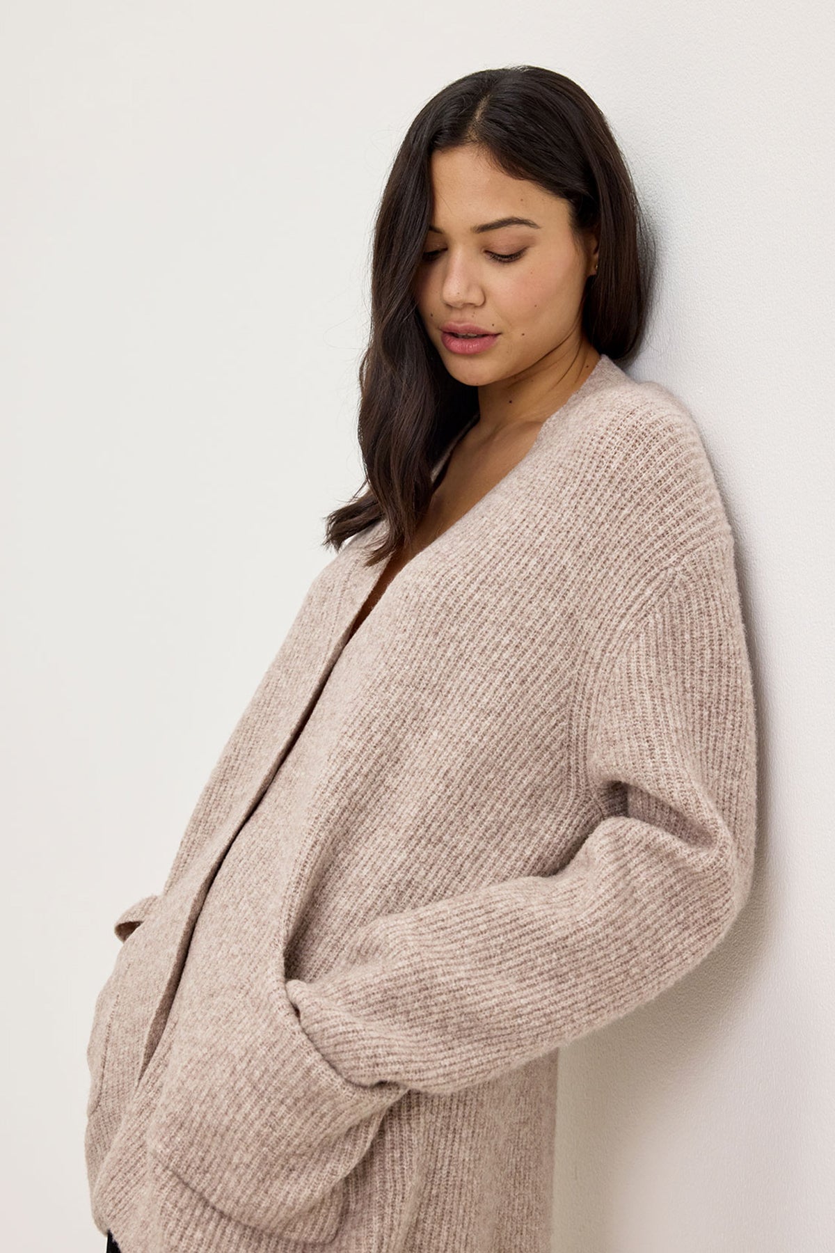 A woman with long hair wears the beige BRITT CARDIGAN by Velvet by Graham & Spencer and stands against a white wall, looking down, embracing the idea of layering for cold weather.-37856069124289