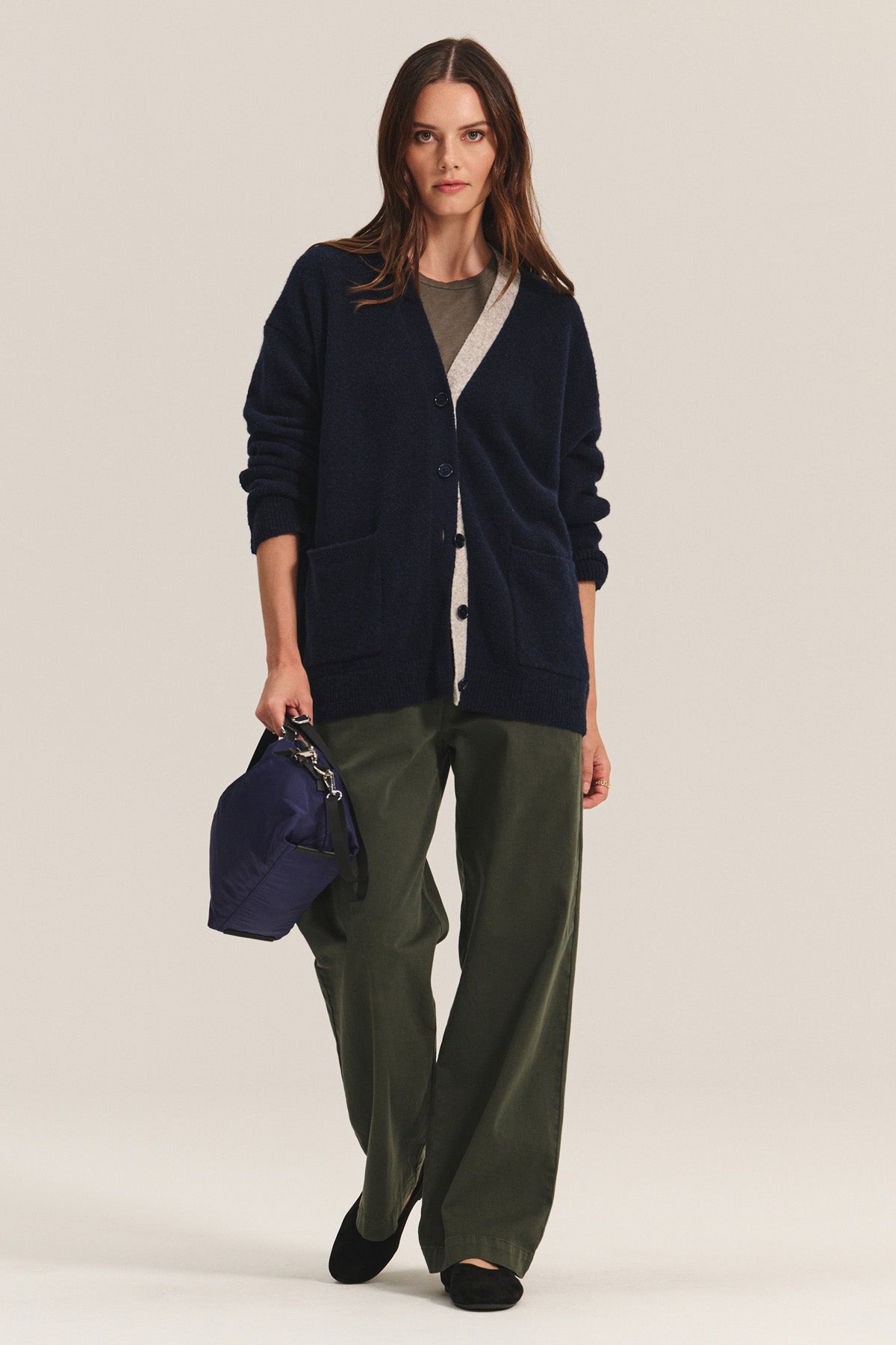 A woman wears the CAMERON CARDIGAN by Velvet by Graham & Spencer over a gray top, paired with olive green wide-leg pants, black shoes, and a navy bag against a plain background.-40333262061761