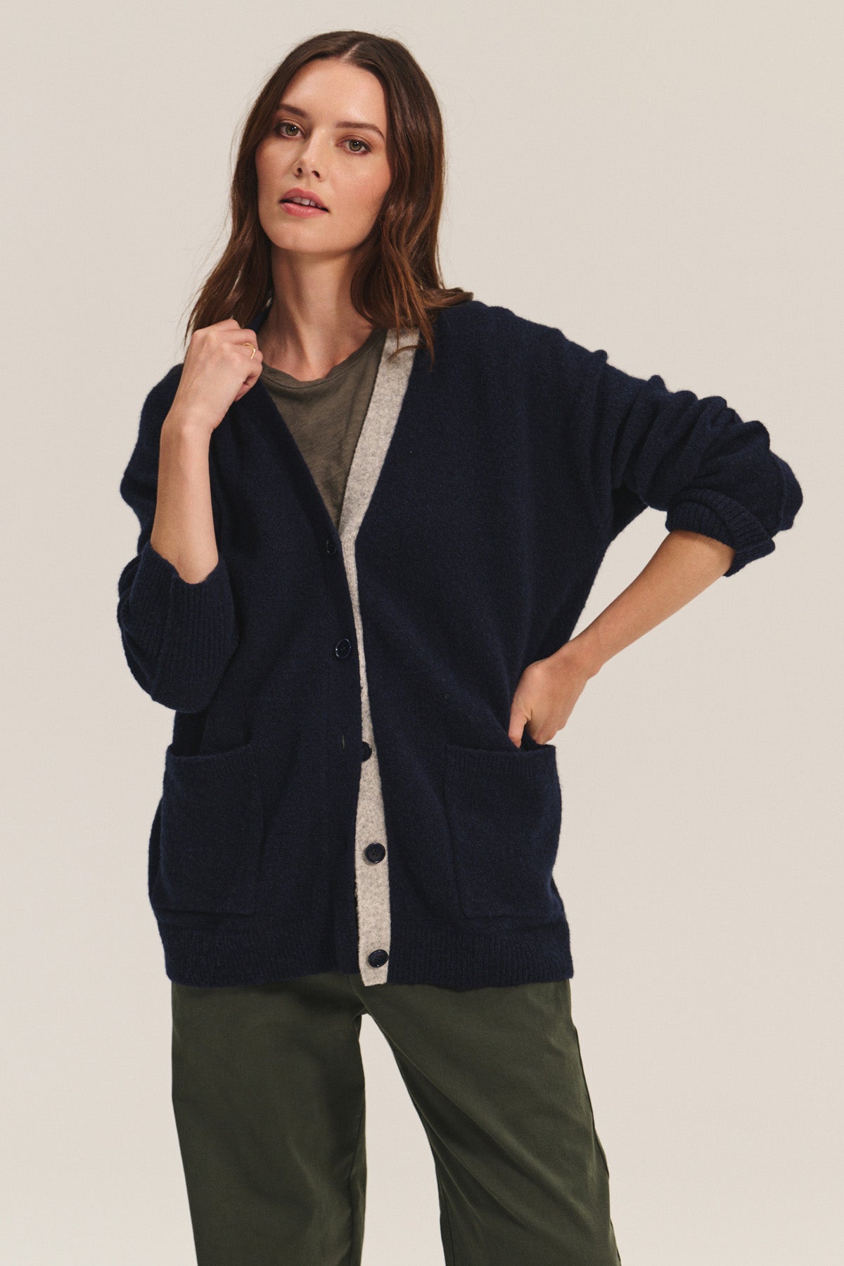 A woman wears the CAMERON CARDIGAN by Velvet by Graham & Spencer—a navy blue wool blend with front pockets—over a gray t-shirt and green pants, standing with one hand in her pocket against a neutral background.