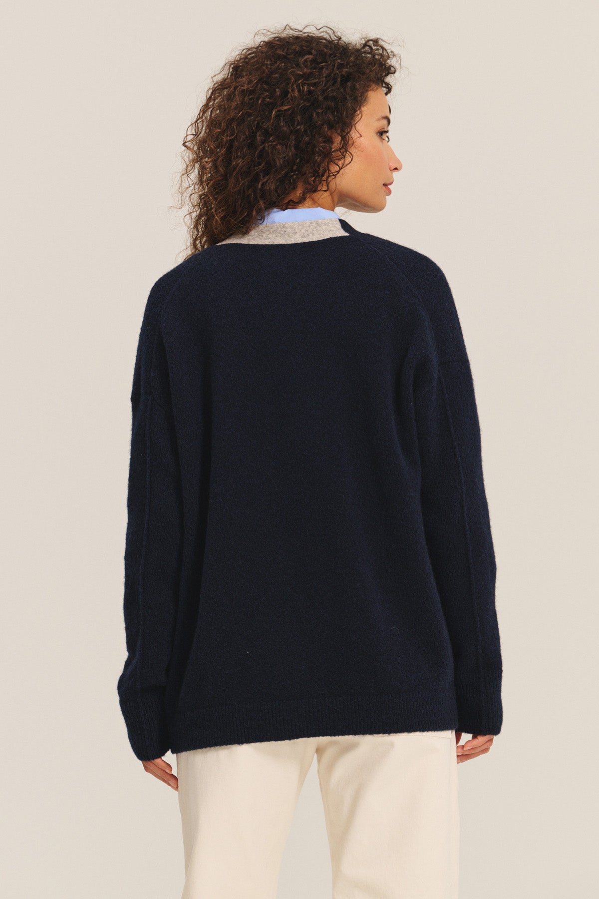 A person with curly hair is seen from the back wearing Velvet by Graham & Spencer's CAMERON CARDIGAN in navy blue with light pants, set against a plain light background.