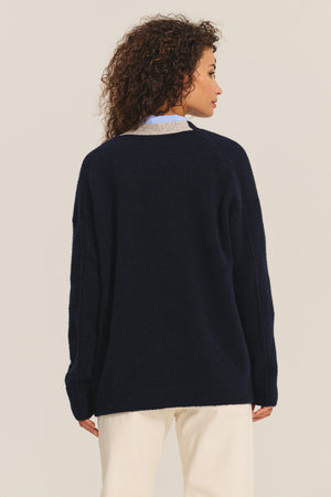 A person with curly hair is seen from the back wearing Velvet by Graham & Spencer's CAMERON CARDIGAN in navy blue with light pants, set against a plain light background.