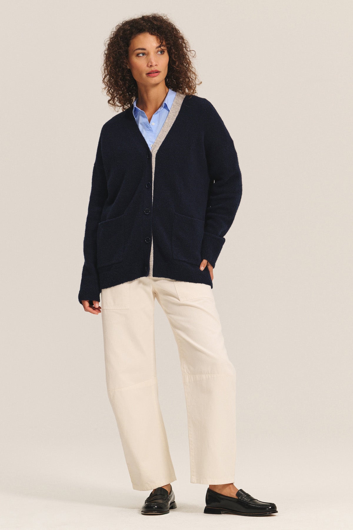 A person wearing a navy cardigan with beige trim, a light blue button-up shirt, Velvet by Graham & Spencer’s BRYLIE SANDED TWILL UTILITY PANT in cream, and black loafers stands against a plain background.-40333257507009