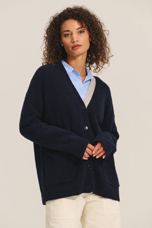 A woman with curly hair wears the CAMERON CARDIGAN by Velvet by Graham & Spencer, a relaxed fit navy blue wool blend cardigan, over a light blue collared shirt and cream pants, standing against a plain background.
