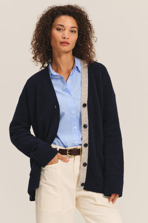 A woman wears the CAMERON CARDIGAN by Velvet by Graham & Spencer, a relaxed-fit wool blend with gray trim, over a light blue button-up shirt and cream pants, standing with one hand in her pocket.