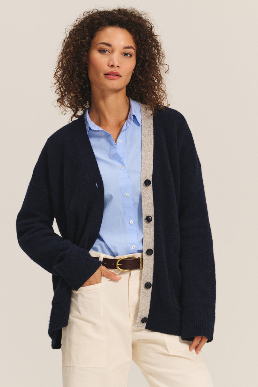 A woman wears the CAMERON CARDIGAN by Velvet by Graham & Spencer, a relaxed-fit wool blend with gray trim, over a light blue button-up shirt and cream pants, standing with one hand in her pocket.