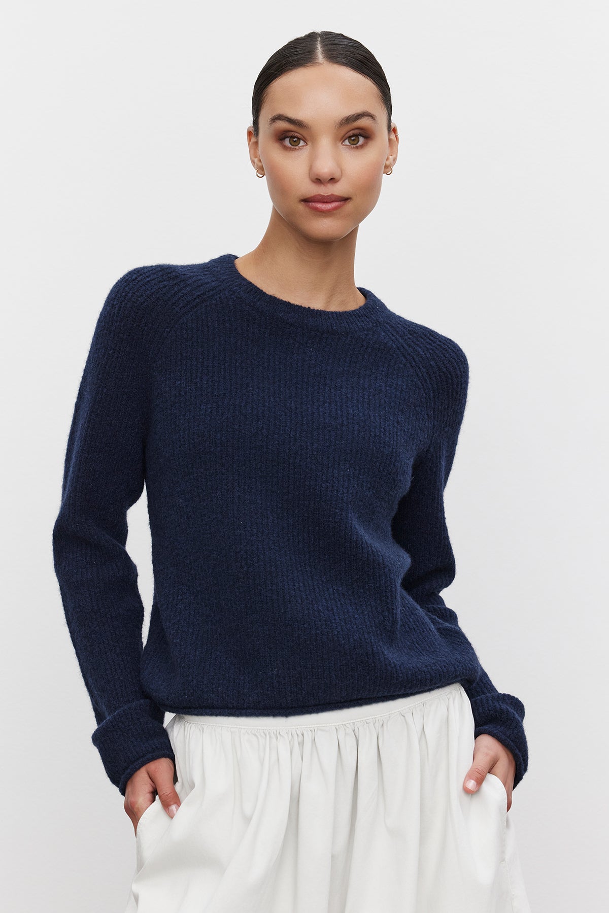   A person with dark hair pulled back, wearing the GIGI SWEATER by Velvet by Graham & Spencer — a cozy knit navy blue sweater with delicate pointelle details — and a white skirt, stands against a plain background. 