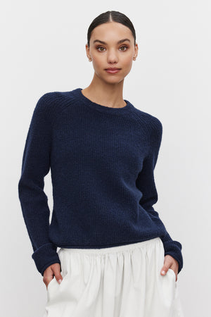 A person with dark hair pulled back, wearing the GIGI SWEATER by Velvet by Graham & Spencer — a cozy knit navy blue sweater with delicate pointelle details — and a white skirt, stands against a plain background.