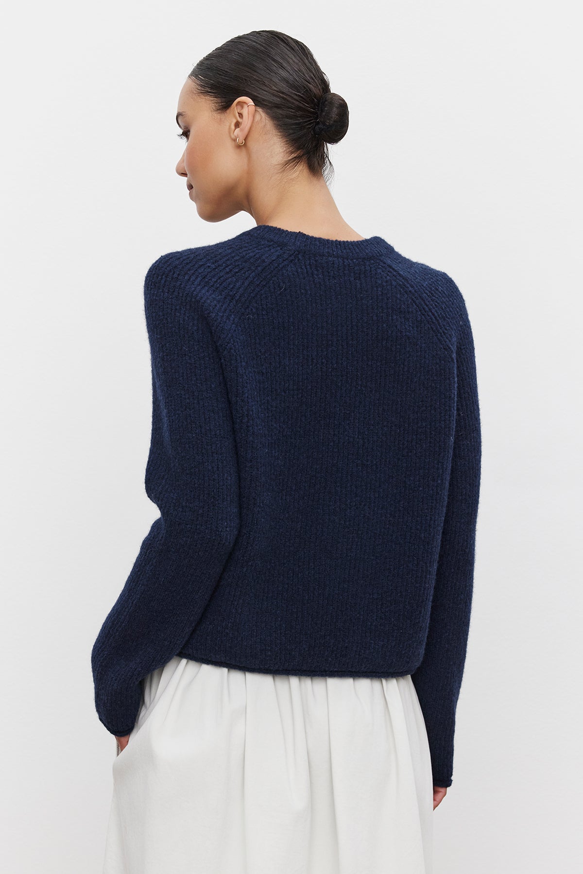   A person with their hair up in a bun is wearing the dark blue GIGI SWEATER by Velvet by Graham & Spencer along with a white skirt, viewed from the back. 