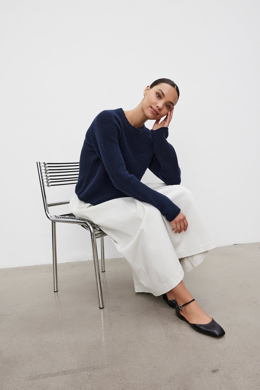 A person wearing the GIGI SWEATER by Velvet by Graham & Spencer, featuring delicate pointelle details, and white wide-leg pants sits on a metal chair with their head resting on their hand. The background is plain white, and the floor is grey.