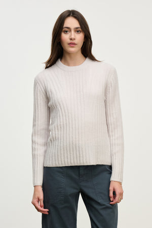 A woman with long brown hair models the IMANA SWEATER by Velvet by Graham & Spencer, a cozy light beige wool blend in ribbed reverse jersey stitch, paired with dark gray pants against a plain light background.