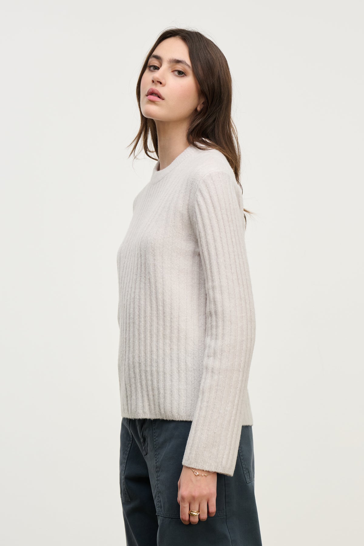 A woman stands sideways against a plain white background wearing the IMANA SWEATER by Velvet by Graham & Spencer in light gray reverse jersey stitch wool blend, paired with dark pants.-40227058581697