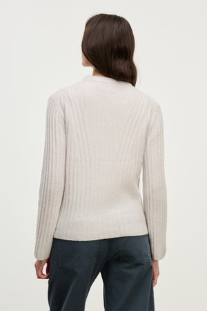 A woman with long brown hair stands with her back to the camera, wearing the IMANA SWEATER by Velvet by Graham & Spencer—a cozy light-colored wool blend with a ribbed knit design—and dark pants against a plain background.