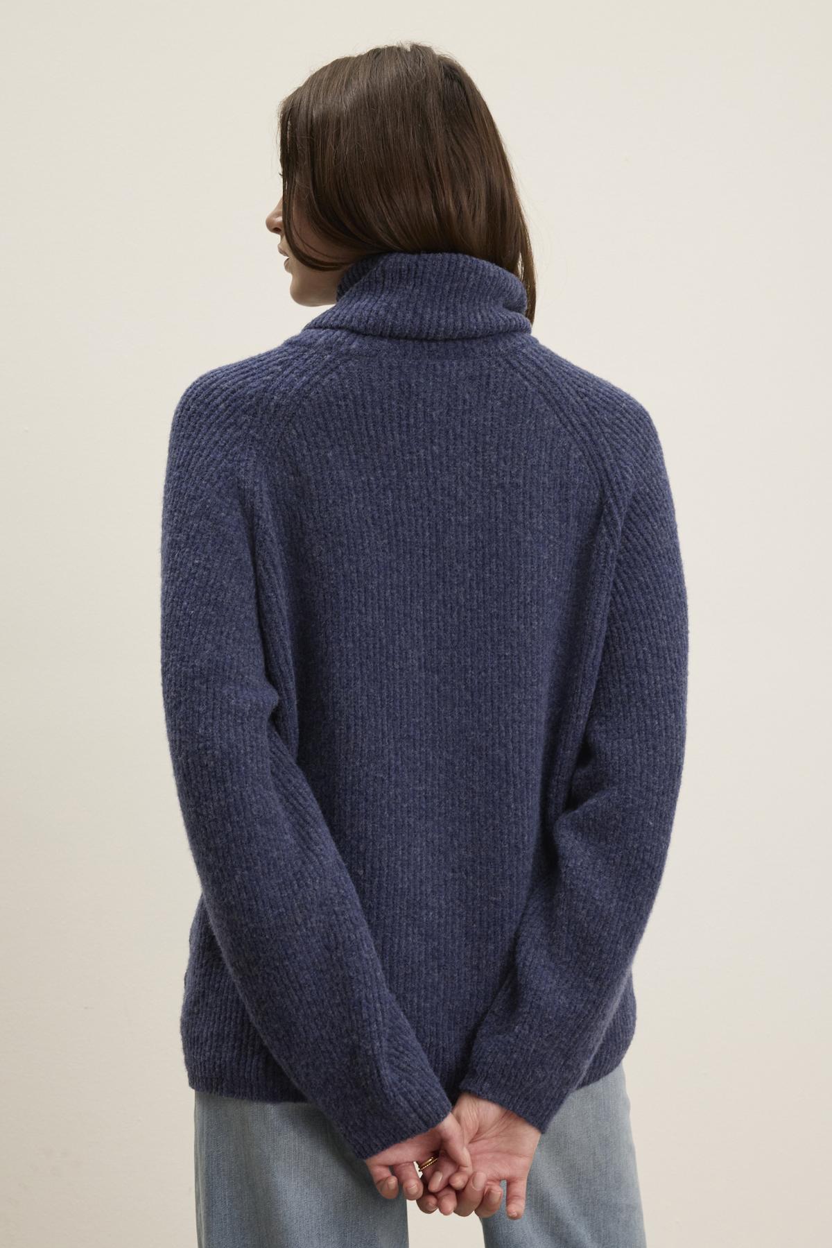   A person with long hair is standing with their back to the camera, wearing a cozy blue JUDITH TURTLENECK SWEATER from Velvet by Graham & Spencer. Their hands are clasped behind their back. 