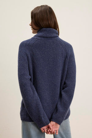 A person with long hair is standing with their back to the camera, wearing a cozy blue JUDITH TURTLENECK SWEATER from Velvet by Graham & Spencer. Their hands are clasped behind their back.