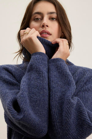 A person wearing the JUDITH TURTLENECK SWEATER by Velvet by Graham & Spencer pulls the cozy wool-blend blue collar close to their face, with their hands positioned near the chin.