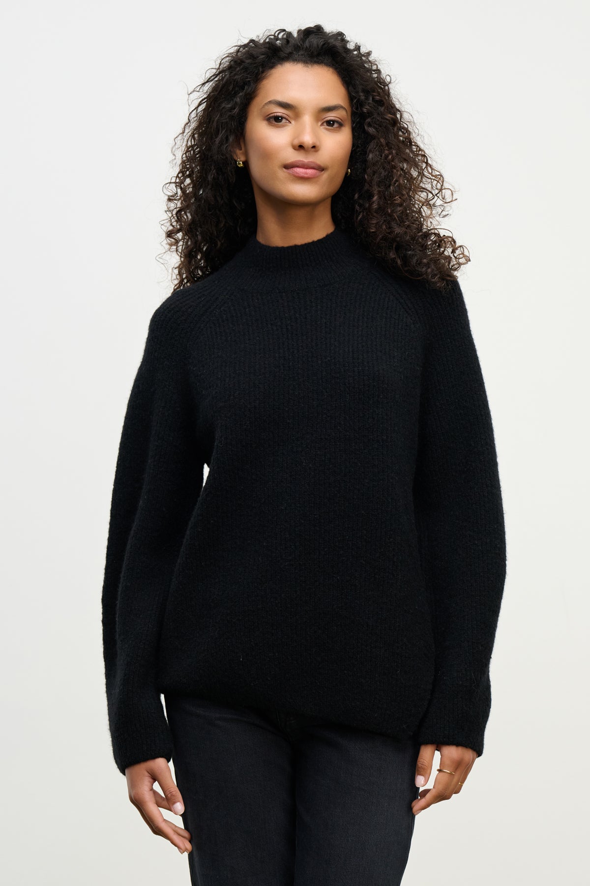 A woman with curly hair wears the black LESA SWEATER by Velvet by Graham & Spencer and black pants, standing against a plain light background.