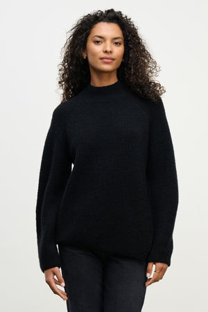 A woman with curly hair wears the black LESA SWEATER by Velvet by Graham & Spencer and black pants, standing against a plain light background.