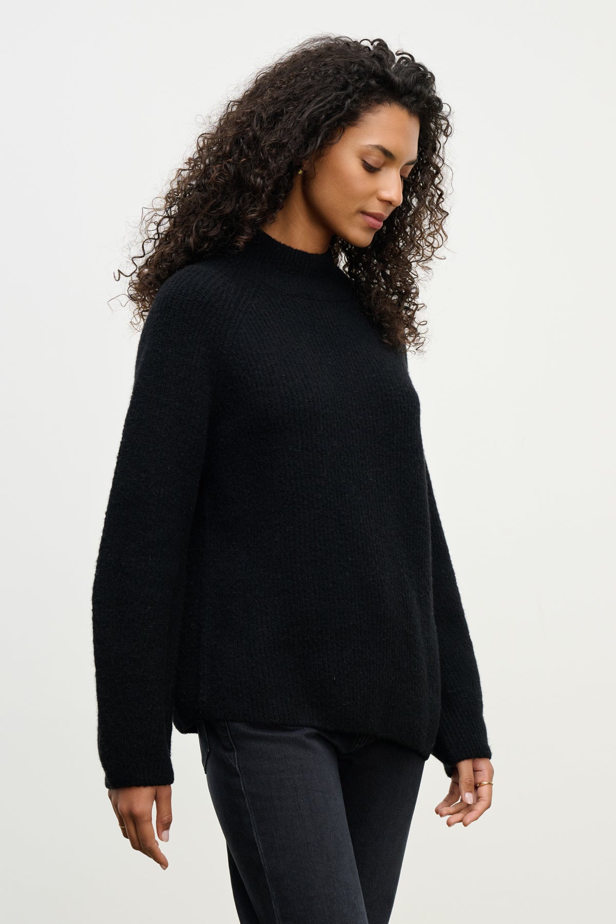 A person with curly hair wears the LESA SWEATER by Velvet by Graham & Spencer in black, paired with dark pants, standing against a light background and looking downward.-40227063005377
