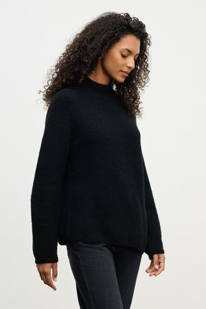 A person with curly hair wears the LESA SWEATER by Velvet by Graham & Spencer in black, paired with dark pants, standing against a light background and looking downward.
