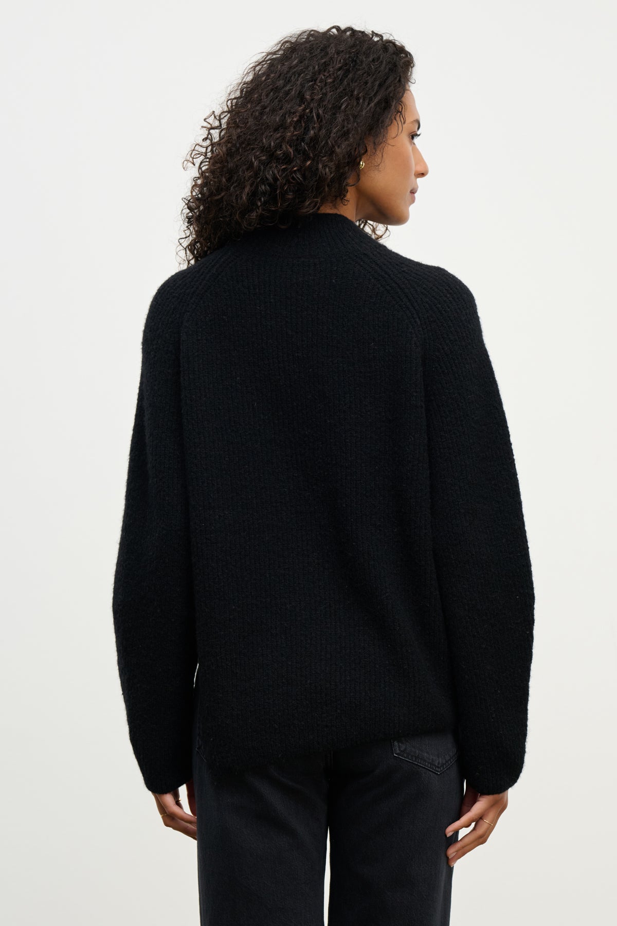 A person with curly hair is shown from the back, wearing the LESA SWEATER by Velvet by Graham & Spencer and black pants against a plain light background.