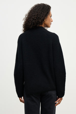 A person with curly hair is shown from the back, wearing the LESA SWEATER by Velvet by Graham & Spencer and black pants against a plain light background.