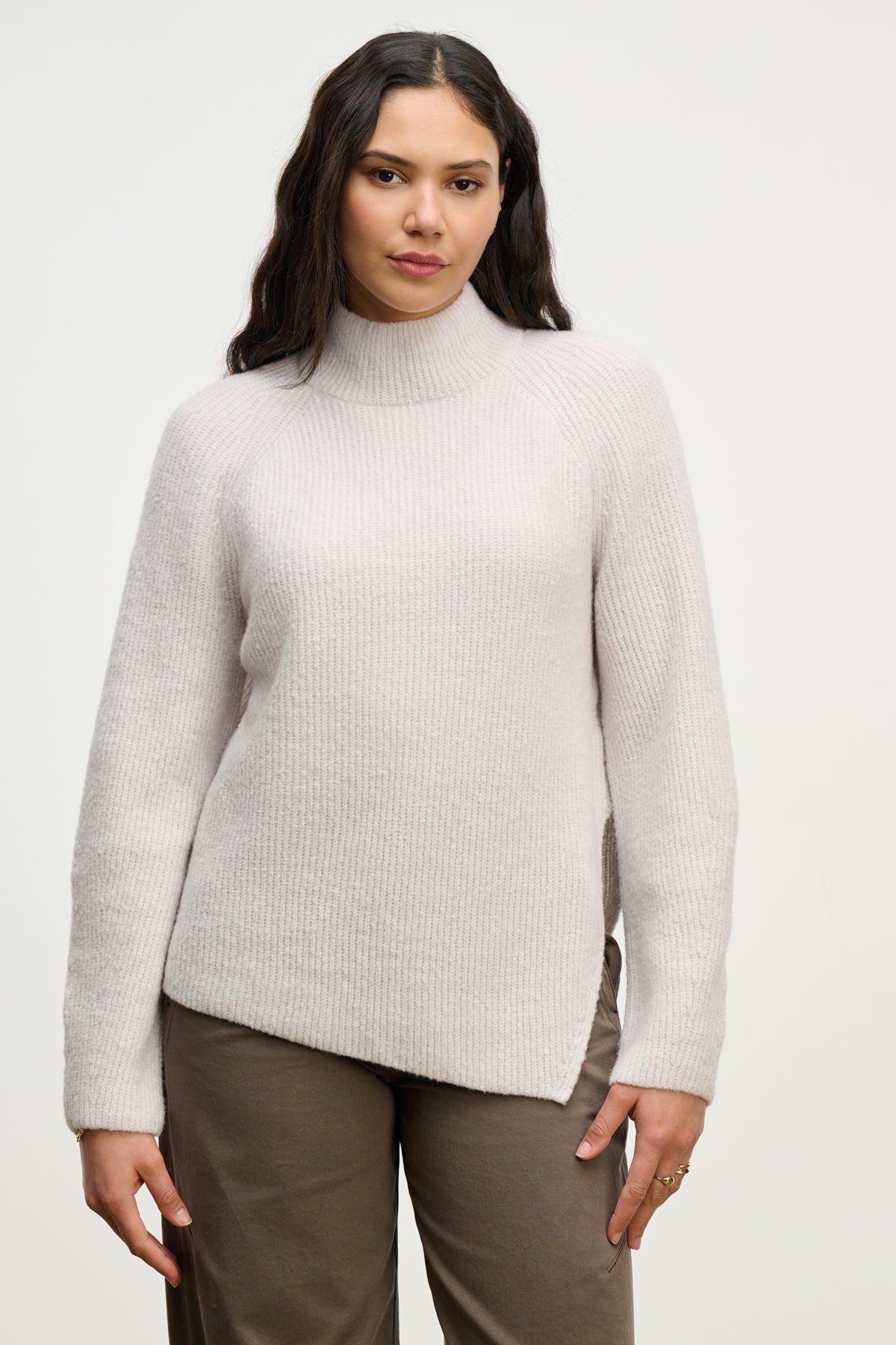   A woman with long dark hair wears the LESA SWEATER by Velvet by Graham & Spencer, a light beige ribbed mock neck style, paired with taupe pants against a plain light background. 