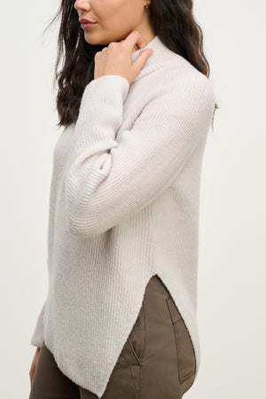 A woman wearing the LESA SWEATER by Velvet by Graham & Spencer—a light beige wool blend with an asymmetrical hem—paired with taupe pants, stands against a plain light background.