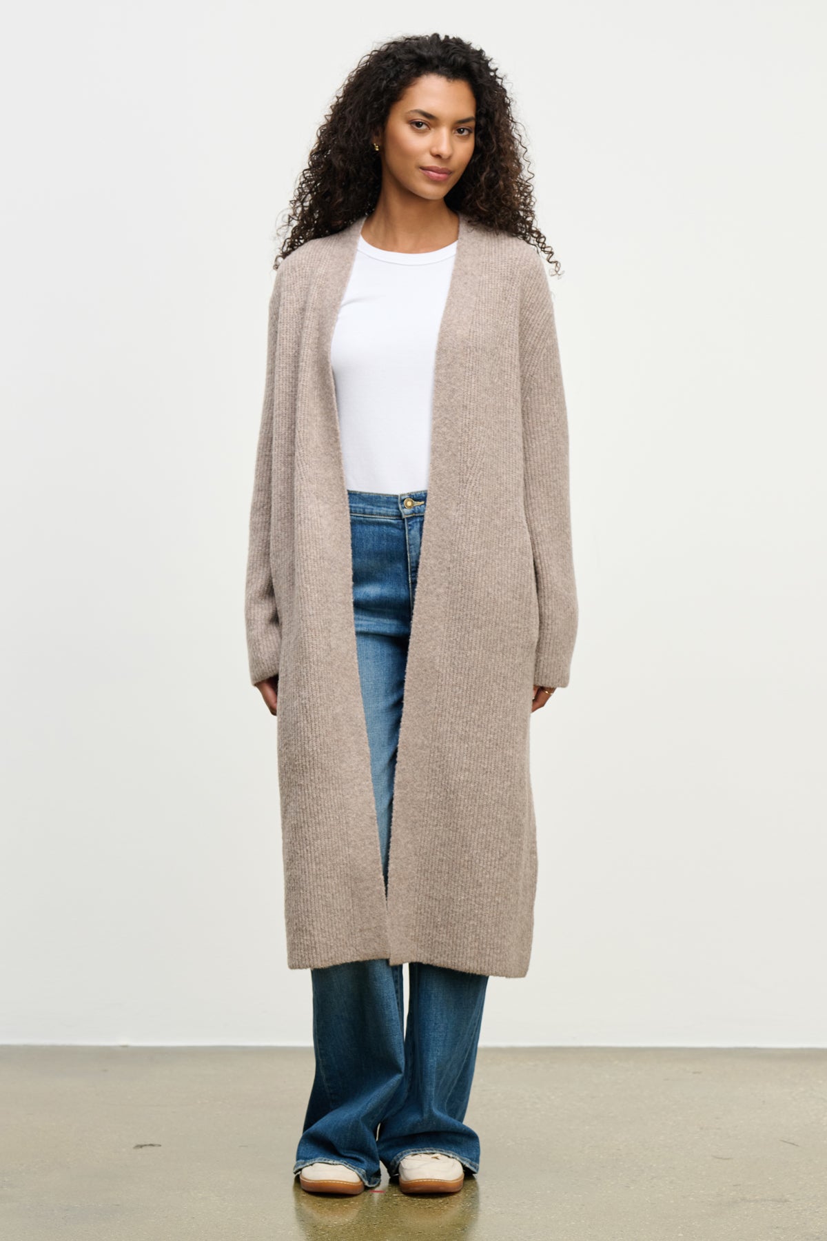   A person stands against a plain background wearing the MISHA CARDIGAN by Velvet by Graham & Spencer—a relaxed, wool blend duster layered over a white shirt, blue jeans, and beige shoes. 
