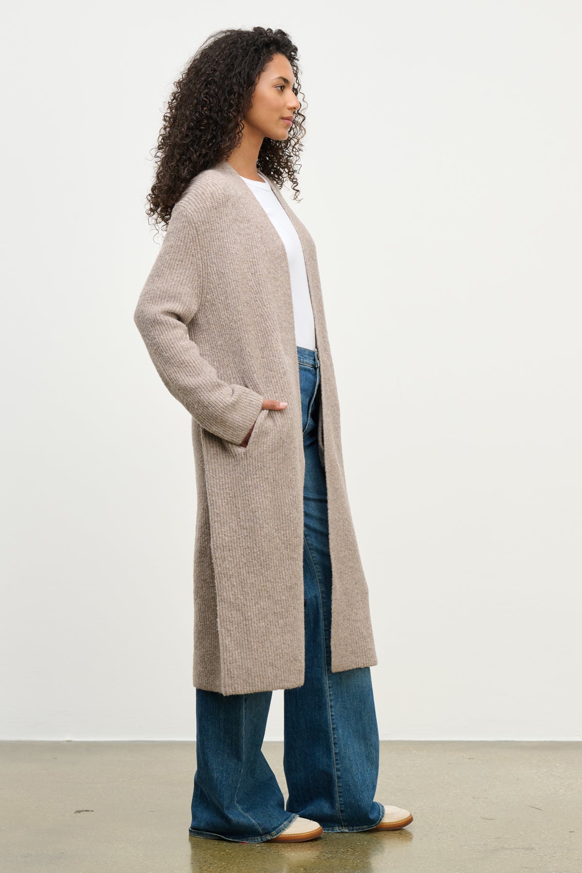 A woman stands in profile against a white wall, wearing the MISHA CARDIGAN by Velvet by Graham & Spencer—an open-front, relaxed-fit style—paired with a white top and wide-leg blue jeans.-40046297514177