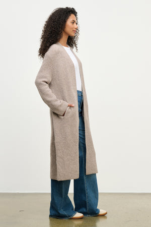 A woman stands in profile against a white wall, wearing the MISHA CARDIGAN by Velvet by Graham & Spencer—an open-front, relaxed-fit style—paired with a white top and wide-leg blue jeans.