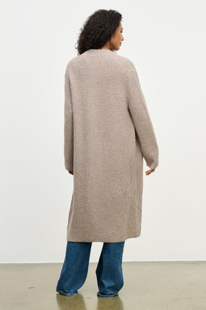 A person with curly hair stands with their back to the camera, wearing the MISHA CARDIGAN by Velvet by Graham & Spencer and blue jeans against a plain light background.