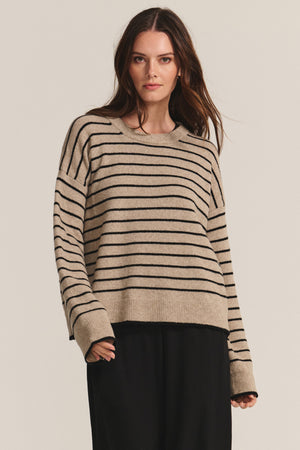 A woman wears the ROMY SWEATER by Velvet by Graham & Spencer—a cozy wool blend with black horizontal stripes—paired with black pants, standing against a plain light background.