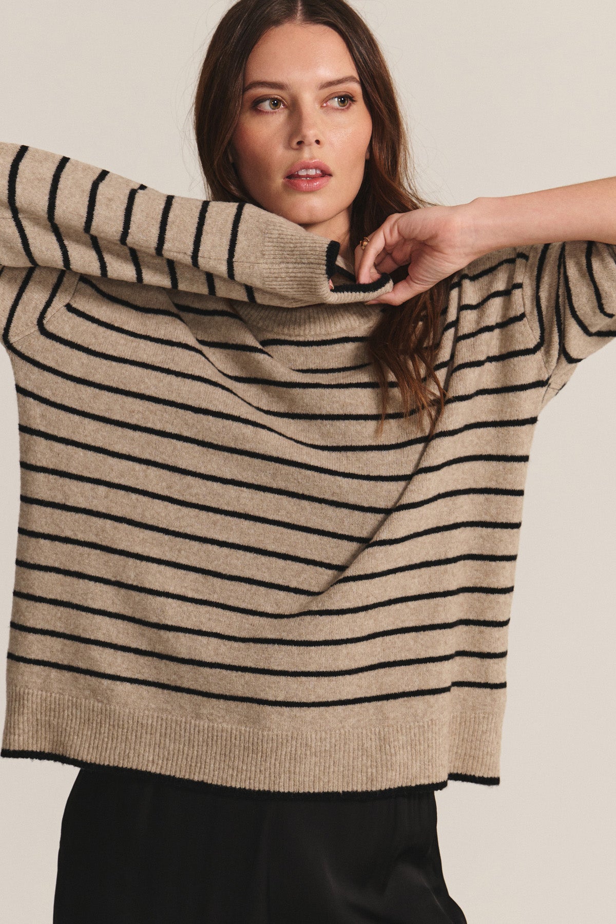 A woman raises her arms and looks to the side while wearing the ROMY SWEATER by Velvet by Graham & Spencer—a beige, relaxed-fit sweater with black stripes—against a plain background.-40672489865409
