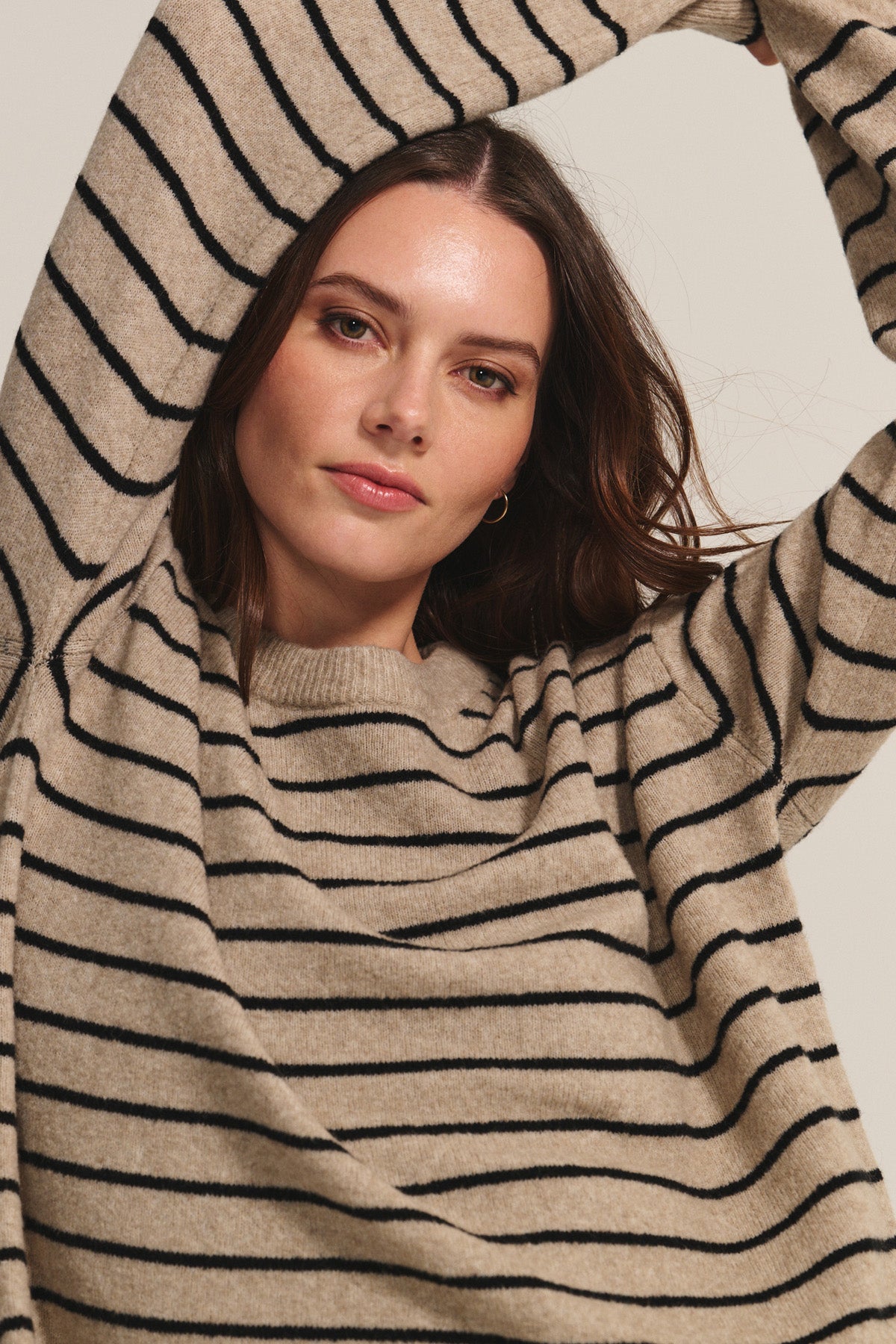 A woman in the Velvet by Graham & Spencer ROMY SWEATER, featuring a cozy relaxed fit and black horizontal stripes, raises her arms above her head and looks directly at the camera.