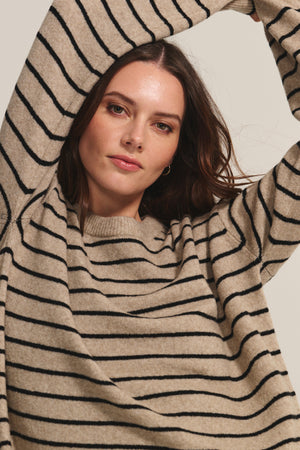 A woman in the Velvet by Graham & Spencer ROMY SWEATER, featuring a cozy relaxed fit and black horizontal stripes, raises her arms above her head and looks directly at the camera.