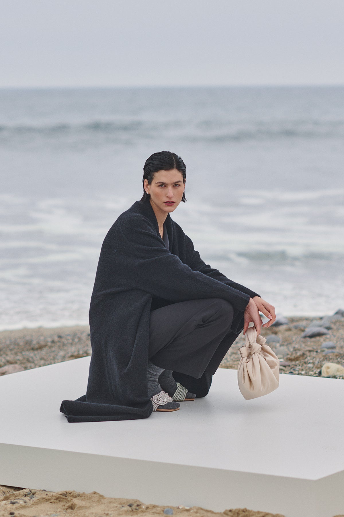 A woman crouching on the beach with a bag, wearing a Velvet by Jenny Graham CARMEL COAT.-35547930460353