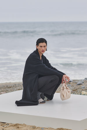 A woman crouching on the beach with a bag, wearing a Velvet by Jenny Graham CARMEL COAT.